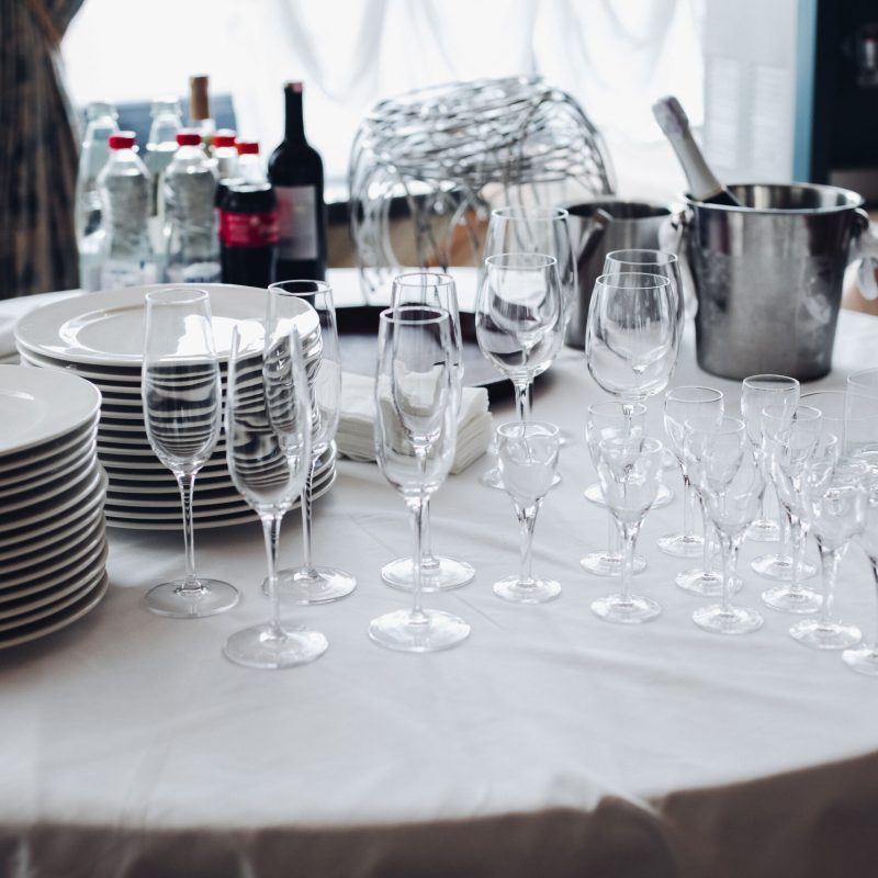 Assortment of kitchenware on white table.View over white round table with stacks of white plates, empty champagne flutes and glasses with bottles of water and ice buckets. View over white round table with stacks of white plates, empty champagne flutes and glasses with bottles of water and ice buckets.